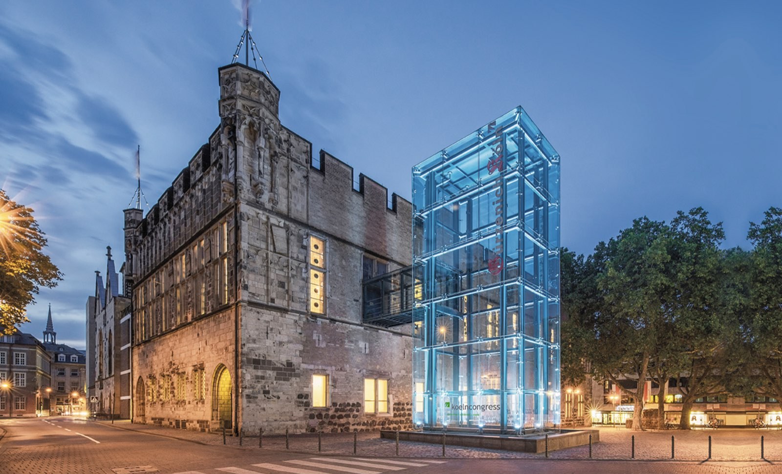 Der Gürzenich in Köln in der Abenddämmerung, daneben der beleuchtete gläserne Aufzugsturm